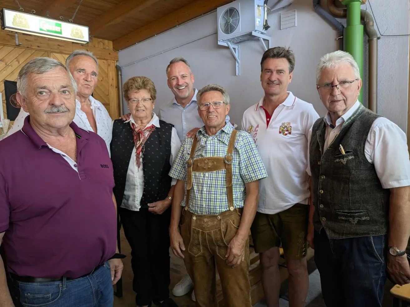 Eine Gruppe von Menschen steht zusammen in einem Raum, lächelt und posiert für ein Foto. Der Mann in der Mitte trägt ein kariertes Hemd und eine Hose mit Aufhängung. Hinter ihm trägt eine Frau einen Schal und eine Brille. Links trägt ein Mann ein lila Hemd. Rechts trägt ein Mann eine Weste.