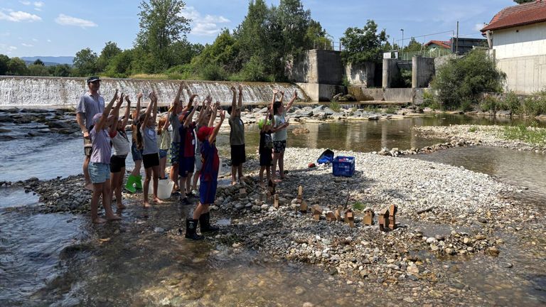 A group of children and adults are standing in a river, raising their hands, and some are wearing boots. There are rocks and a blue crate on the riverbank.