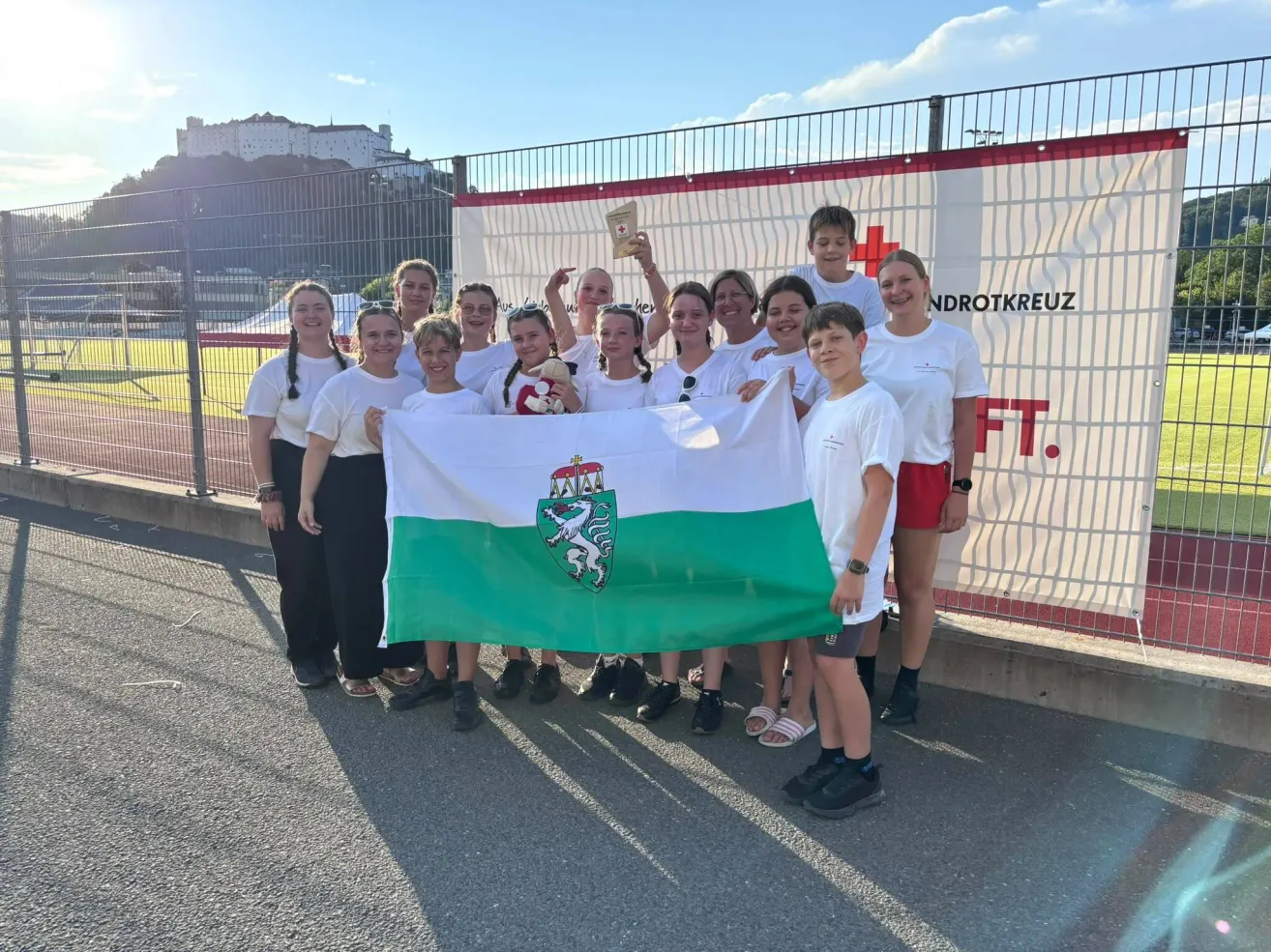 A group of young people in white shirts and shorts hold a flag with a lion emblem, standing in front of a banner with red crosses and text.