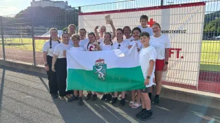 A group of young people in white shirts and shorts hold a flag with a lion emblem, standing in front of a banner with red crosses and text.
