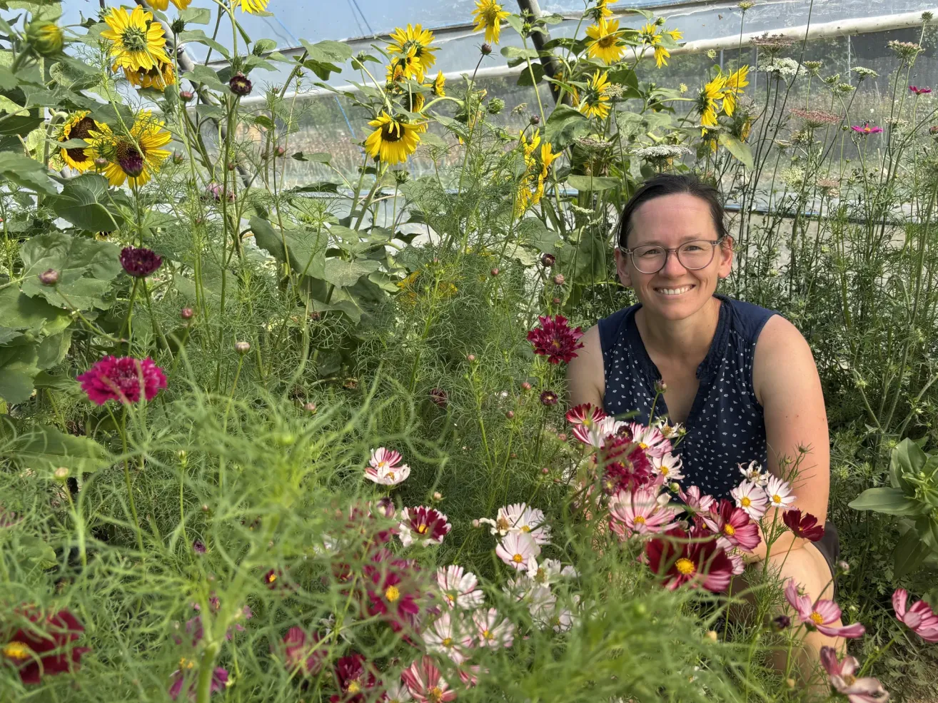 A woman in a greenhouse surrounded by a variety of flowers, including sunflowers and daisies, smiling for the camera.