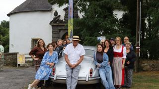 A group of people are posing for a photo in front of a vintage car, with a man in a hat standing in the middle. The background features a building with a banner and a tree.