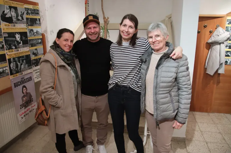 Vier Menschen stehen zusammen und lächeln für ein Foto. Ein Mann in einem schwarzen T-Shirt und eine Frau in einem gestreiften T-Shirt stehen in der Mitte, mit zwei Frauen auf beiden Seiten. Die Frau auf der linken Seite trägt einen beigen Mantel, und die Frau auf der rechten Seite trägt eine graue Jacke.