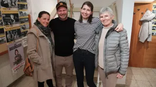 Four people are standing together, smiling for a photo. A man in a black shirt and a woman in a striped shirt are in the middle, with two women on either side. The woman on the left wears a beige coat, and the woman on the right wears a gray jacket.