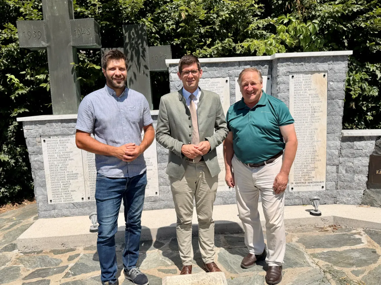 Three men stand in front of a memorial wall with plaques, smiling and posing for a photo. The man on the left wears a light blue shirt and jeans, the middle man a light green suit with a tie, and the man on the right a green shirt and khaki pants.