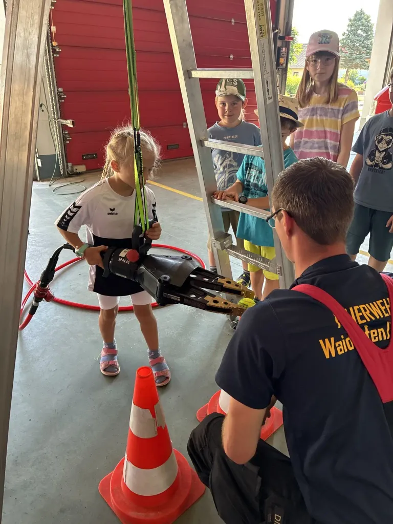 A young girl in a white shirt and shorts is holding a mechanical arm while a man in a blue shirt and red suspenders is assisting her. Behind them, several children are standing near a ladder and a red fire extinguisher.