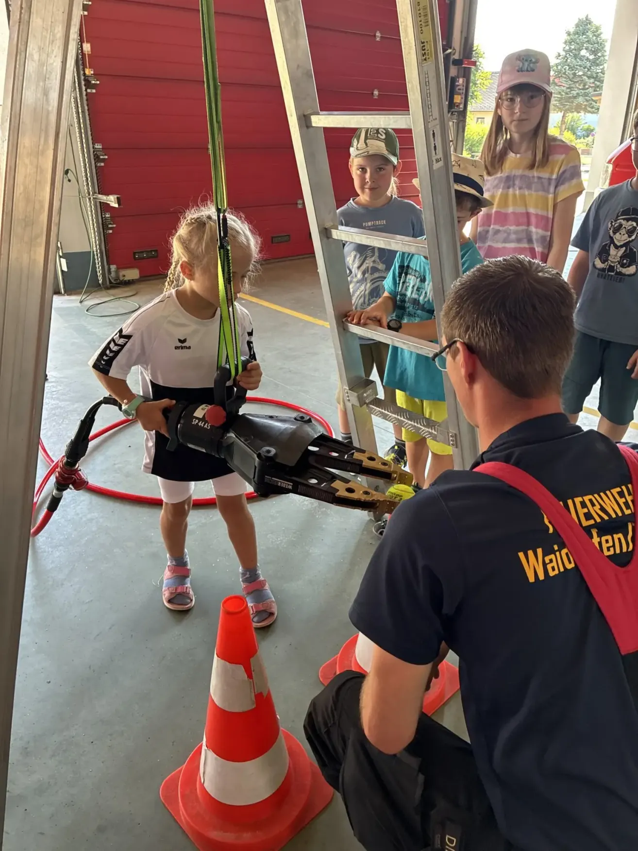 A young girl in a white shirt and shorts is holding a mechanical arm while a man in a blue shirt and red suspenders is assisting her. Behind them, several children are standing near a ladder and a red fire extinguisher.