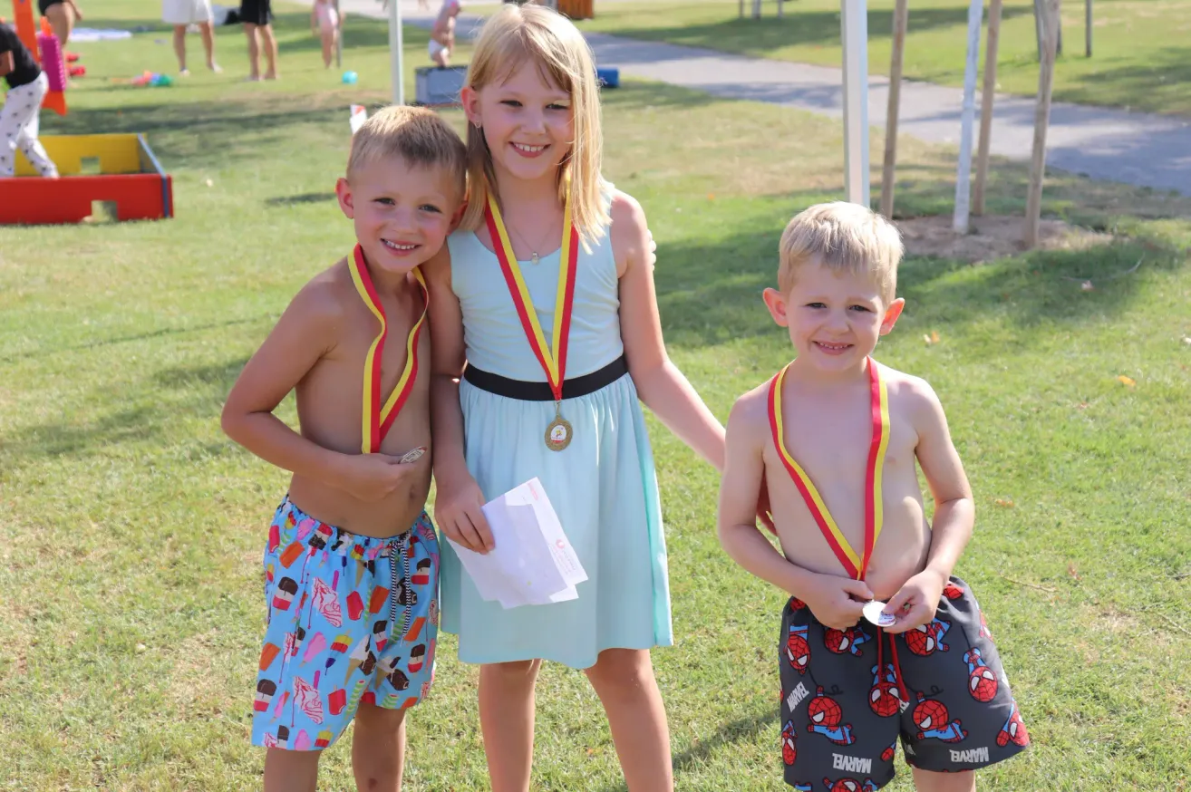 Three children, two boys and a girl, stand together on a grassy field wearing medals. The girl holds a certificate and smiles. The boys wear swim trunks with colorful prints.