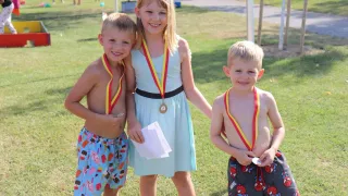 Three children, two boys and a girl, stand together on a grassy field wearing medals. The girl holds a certificate and smiles. The boys wear swim trunks with colorful prints.