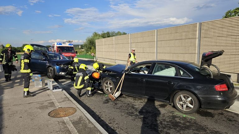 Emergency responders in yellow vests work on a damaged black car on the roadside. A fire truck is parked behind a barrier.