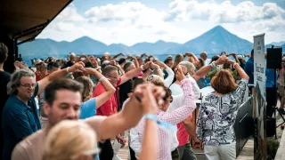A group of people are dancing outdoors, with mountains in the background. Some are wearing bracelets and glasses. The sky is blue with clouds.