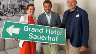 Drei Personen stehen in einem Raum und halten ein Schild mit der Aufschrift 'Grand Hotel Sauerhof'. Die Frau links trägt ein orangefarbenes Kleid und die beiden Männer tragen Anzüge.