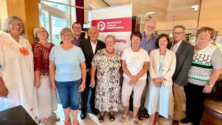 A group of people, both men and women, are standing together in a room, smiling for a photo. They are wearing casual clothes and some are wearing glasses. Behind them is a banner with the text 'PENSIONISTEN VERBAND OBEROSTERREICH'.