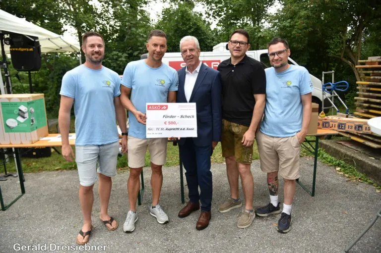 Five men stand together holding a check for 500 Euros. They are dressed casually and appear to be posing for a photo.