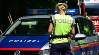 Eine weibliche Polizeibeamtin steht neben einem Polizeiwagen mit blinkenden Lichtern auf dem Dach. Sie trägt eine reflektierende Weste mit dem Wort 'POLIZEI'.