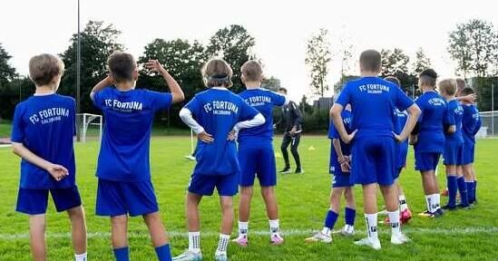 A group of young soccer players in blue uniforms with FC Fortuna on their backs are standing on a grassy field, possibly getting ready for a game.