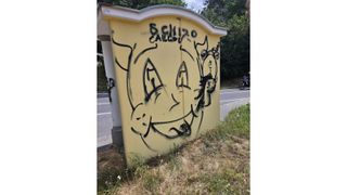 A yellow structure with graffiti on it, featuring a smiley face and the word 'SCH120'. It is located on the side of a road with a motorcycle in the background.