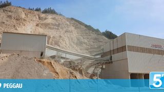 A mining facility with a conveyor belt transporting materials from a large pile of dirt to a processing plant. The facility is situated on a steep hill with a clear blue sky in the background.