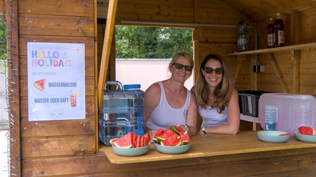 Zwei Frauen mit Sonnenbrillen und weißen Tanktops sitzen hinter einem Tresen in einer hölzernen Kabine, lächeln und posieren für ein Foto. Vor ihnen stehen zwei Schüsseln mit Wassermelonenscheiben. Dahinter steht ein Wasserkrug und ein Schild an der Wand.