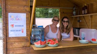 Two women wearing sunglasses and white tank tops are sitting behind a counter in a wooden booth, smiling and posing for a photo. In front of them are two bowls of watermelon slices. Behind them is a water jug and a sign on the wall.