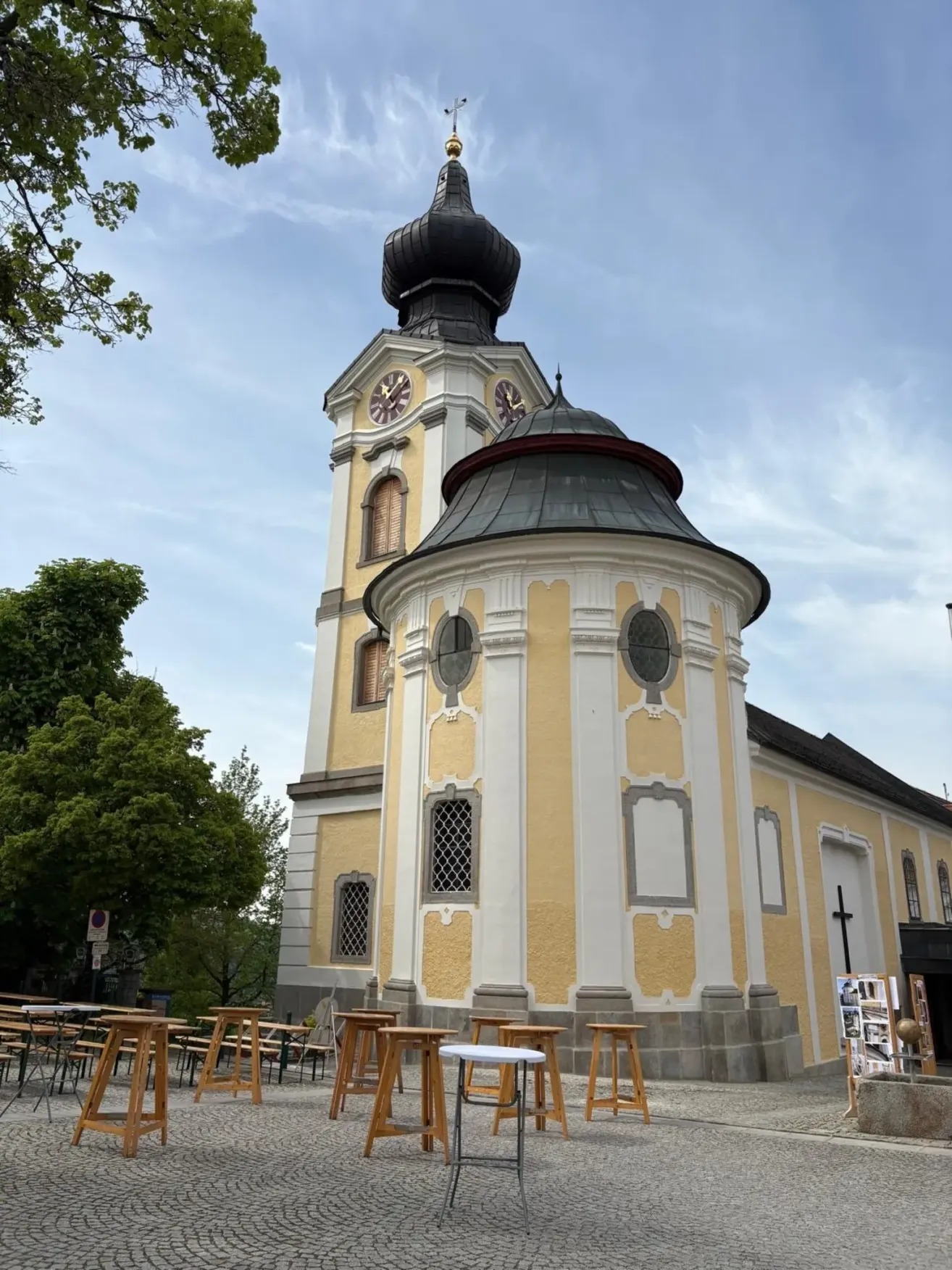 Eine gelbe und weiße Kirche mit einer schwarzen Kuppel und einem Uhrturm, umgeben von Holztischen und Stühlen unter einem blauen Himmel.