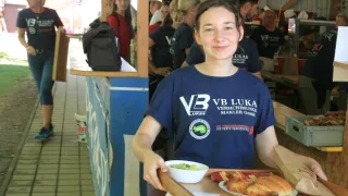 A woman wearing a blue shirt with the text 'VB LUKAS' is smiling and holding a tray with food. Behind her, several people are standing and talking. A black bag is on the counter.