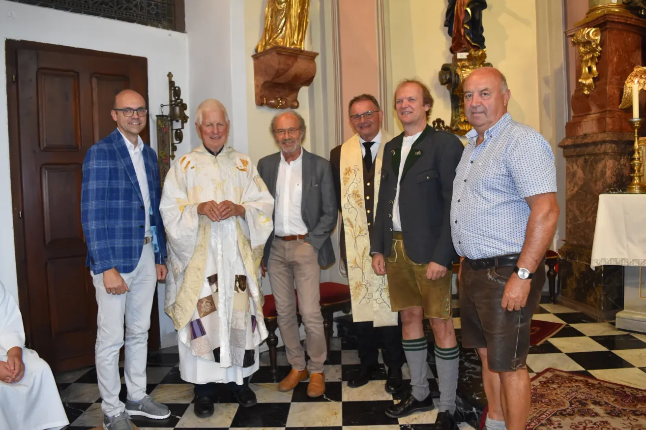 Five men stand in a church, one wearing a white robe, another in a brown vest, and the rest in casual attire, smiling for a photo.