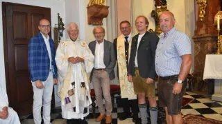 Five men stand in a church, one wearing a white robe, another in a brown vest, and the rest in casual attire, smiling for a photo.