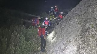 A group of hikers in red jackets and helmets are climbing up a rocky mountain at night. They are equipped with headlamps and appear to be navigating a challenging path.