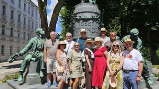 A group of tourists poses for a photo in front of a statue of Josef Wendl in a park. The statue is surrounded by greenery and two smaller statues. The tourists are wearing hats and sunglasses.