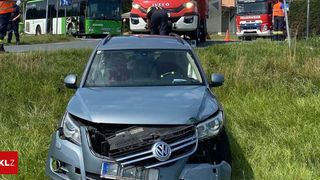 A damaged silver Volkswagen SUV is parked on the side of a road with emergency vehicles nearby. A person in uniform is inspecting the vehicle.