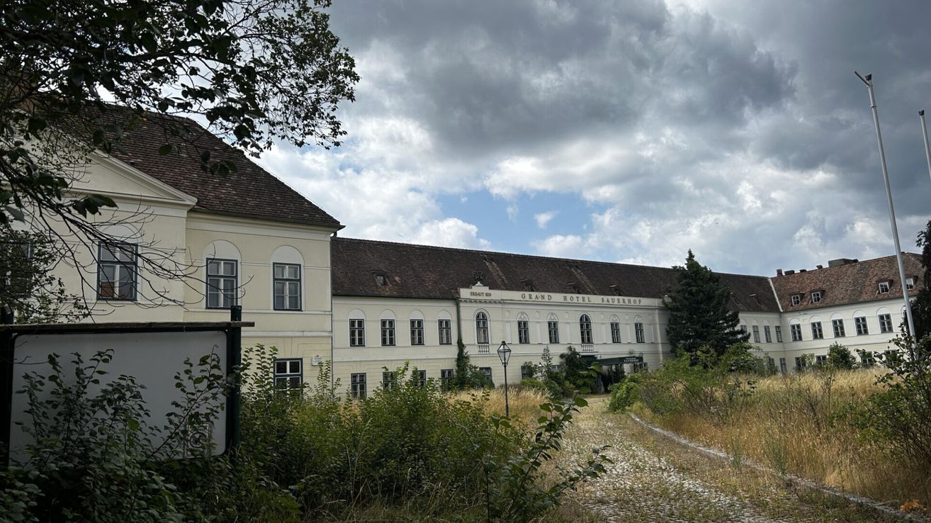 An old, large building with a brown roof and many windows, labeled Grand Hotel Sauerhof, stands in a field with overgrown grass and bushes.
