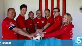A group of men and a woman in red shirts are holding a soccer ball together in a room with a red and white striped wall.