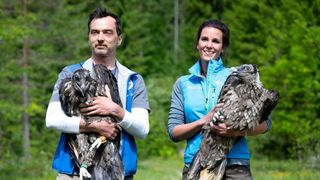 A man and a woman are holding a bird with spread wings in their hands, smiling and posing for a photo.