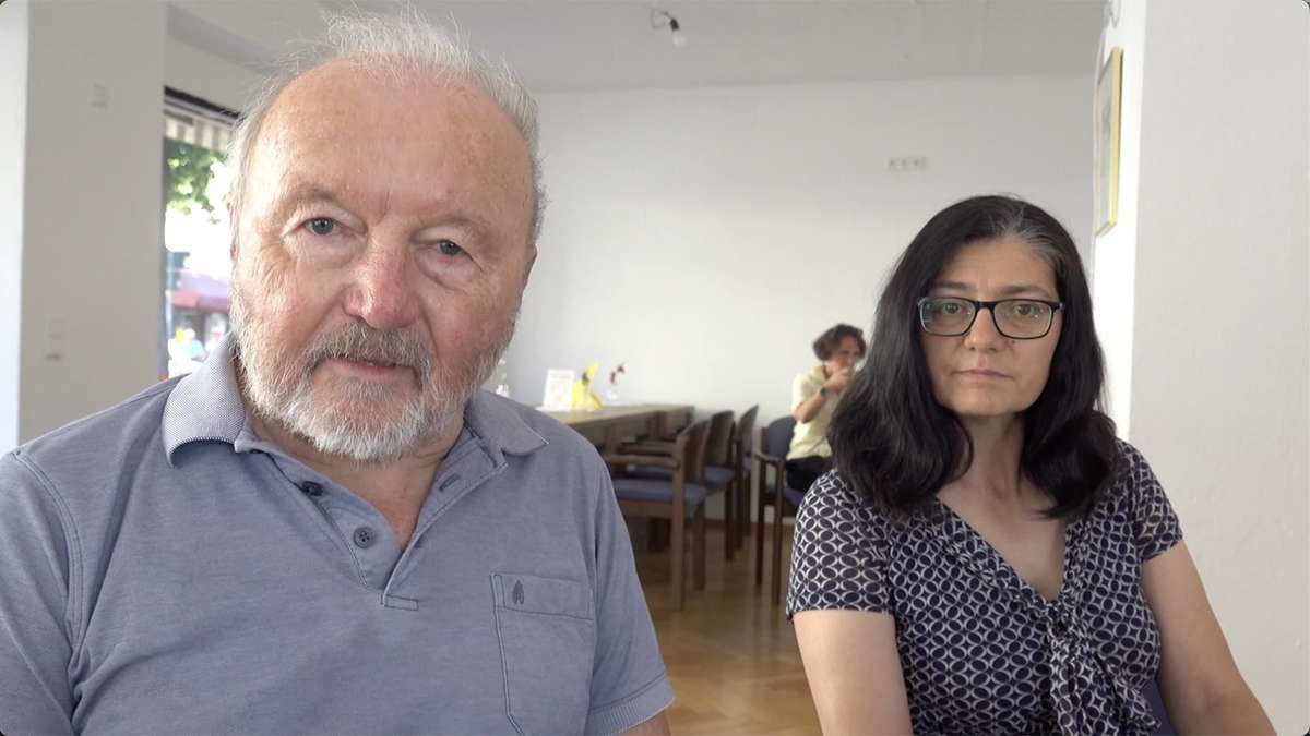 An older man with a white beard and mustache is standing next to a woman wearing glasses. They are both looking at the camera. Behind them, there is a table with chairs and a woman sitting. The room has white walls and wooden floors.