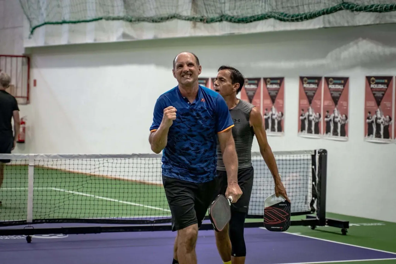 Zwei Männer stehen auf einem Paddle-Tennisplatz, einer hält eine Paddel und der andere lächelt. Sie tragen beide Sportbekleidung. Hinter ihnen ist ein Netz und eine Wand mit Postern.