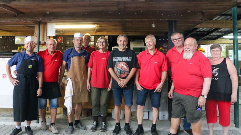 A group of adults in red shirts and shorts, one wearing a chef's hat and apron, standing in a room with wooden beams and a bright light.
