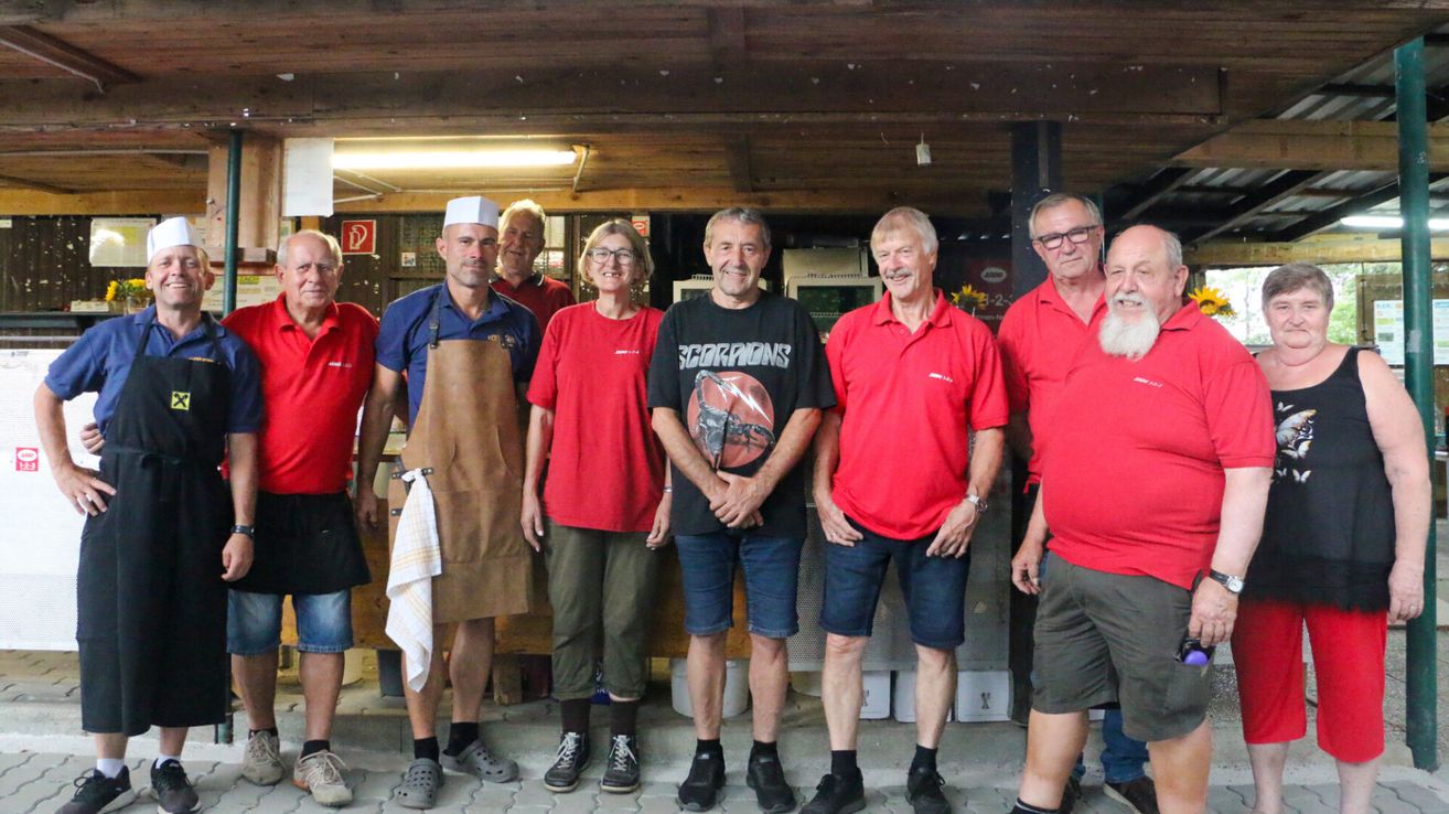 A group of adults in red shirts and shorts, one wearing a chef's hat and apron, standing in a room with wooden beams and a bright light.