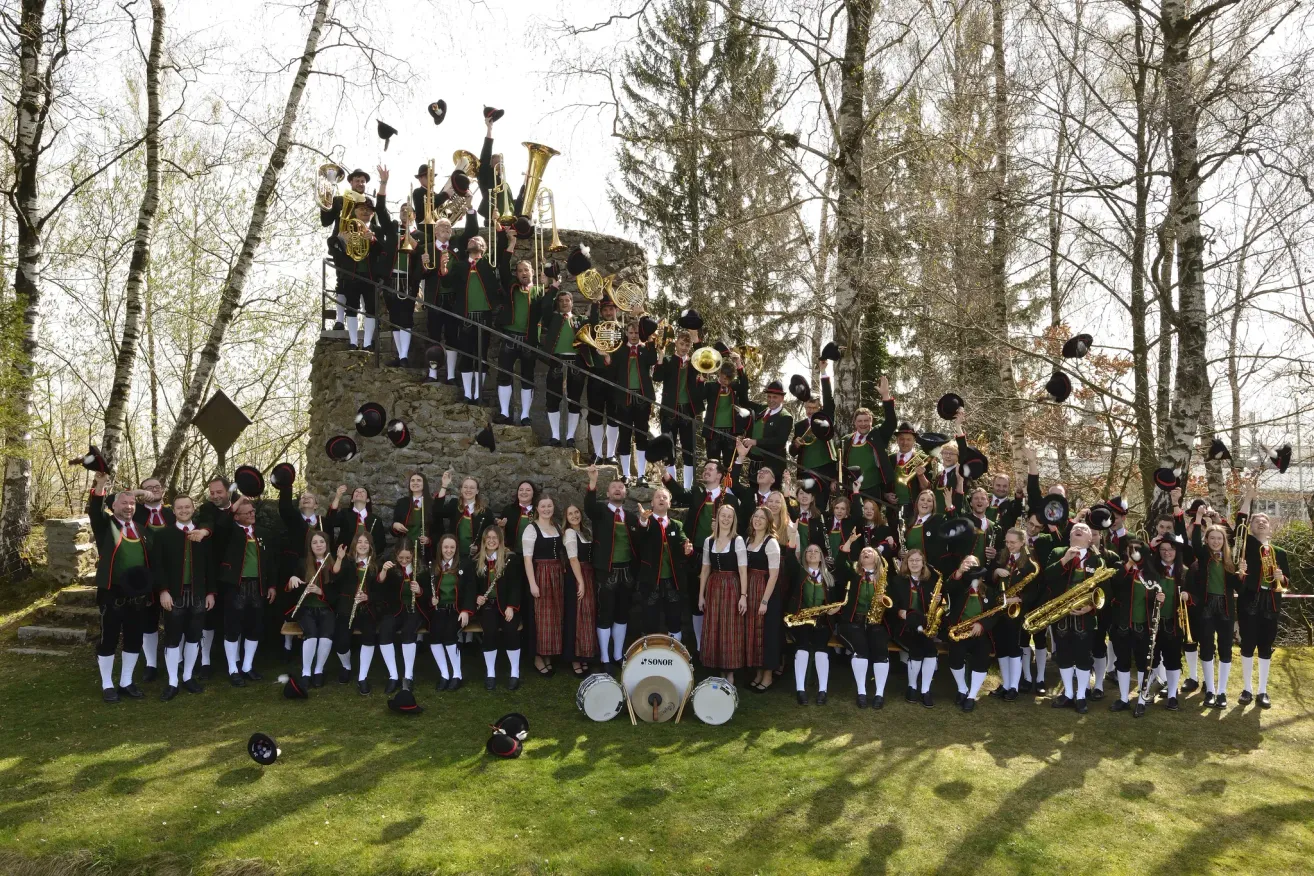 A large group of musicians in green and white uniforms, some standing on a stone wall, others on the grass, all holding instruments and hats.