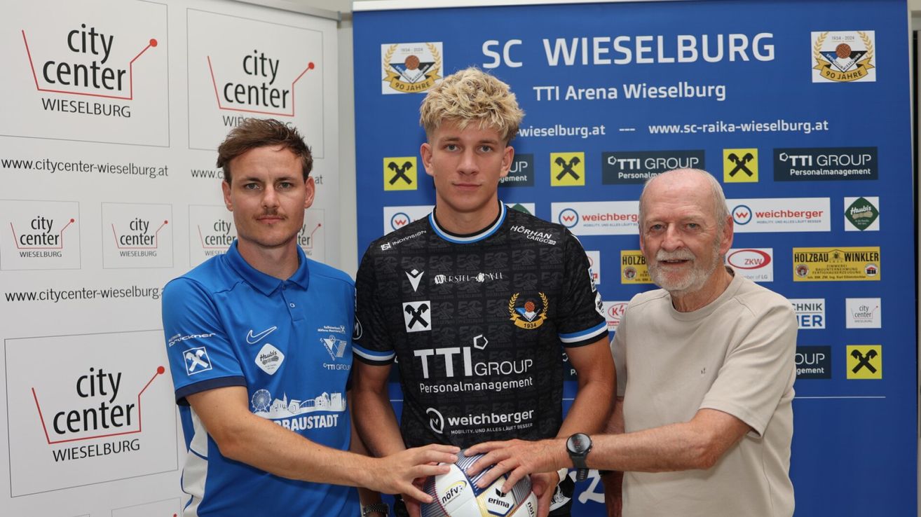 Three men stand in front of a blue backdrop with logos and text. The man in the middle holds a soccer ball, while the man on the left wears a blue shirt and the man on the right wears a white shirt.
