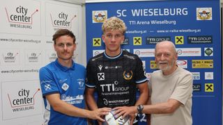Three men stand in front of a blue backdrop with logos and text. The man in the middle holds a soccer ball, while the man on the left wears a blue shirt and the man on the right wears a white shirt.