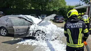A firefighter in a yellow helmet stands near a wrecked car, spraying foam from a hose. The car is covered in foam and is parked on a paved area.