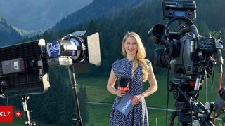 A woman with blonde hair stands in front of a camera with a microphone, holding a sign that reads Studio. Behind her, there are two cameras on tripods, and a mountain landscape is visible in the background.