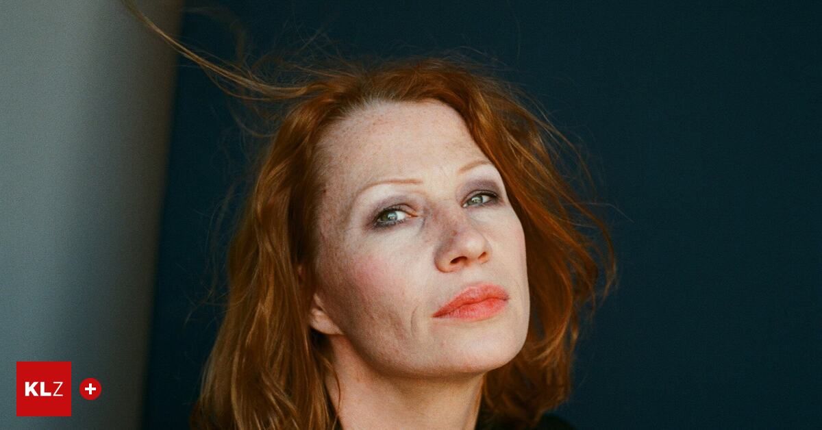 A close-up portrait of a woman with red hair and green eyes, her face slightly tilted, and her hair blowing in the wind against a dark background.