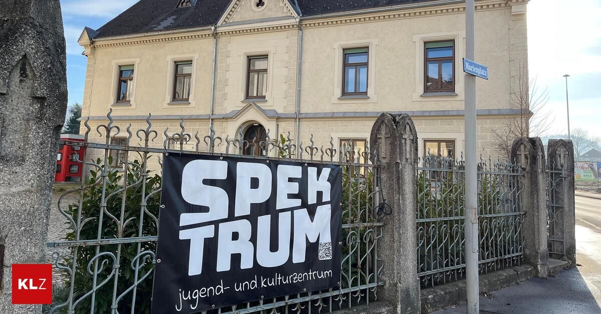 A banner with the text 'Spek Trum' is hanging on a fence in front of a building with a black roof and several windows.