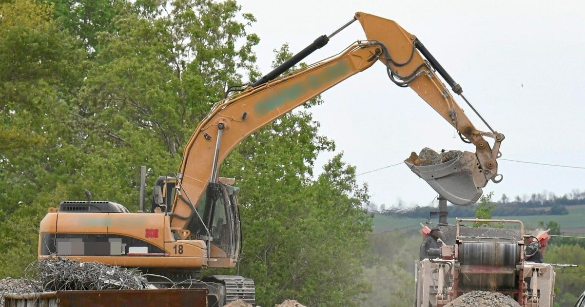 A yellow excavator with the number 18 on its front is dumping debris into a truck on a construction site.
