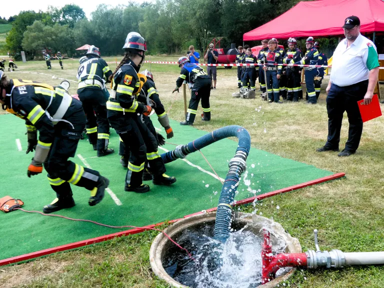 Firefighters in full gear are practicing a water rescue operation on a green mat, with water spraying from a hose into a manhole.