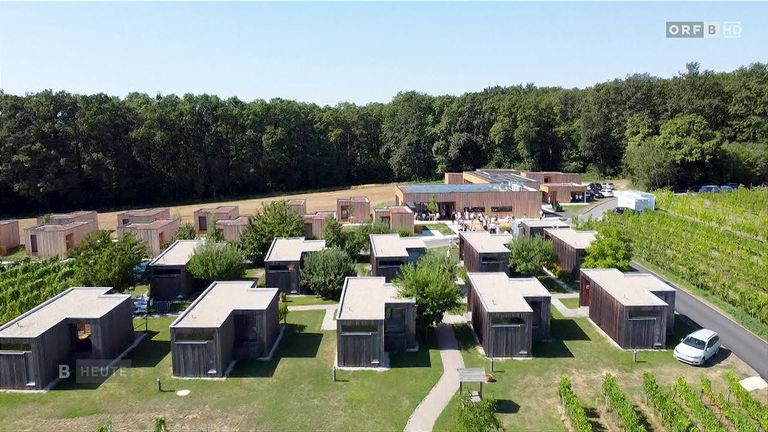 Luftaufnahme eines modernen architektonischen Komplexes mit mehreren kleinen Holzhäusern und einem großen Gebäude im Hintergrund, umgeben von Grün.