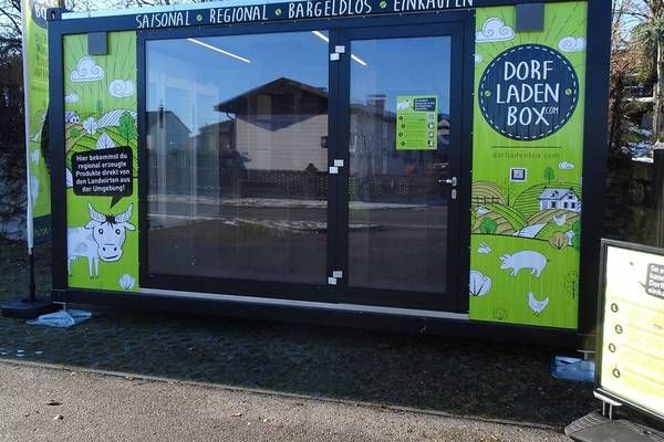 A green and black mobile store with glass doors and windows, featuring a cow and a house on the side. The store has a sign that reads Dorf Laden Box.