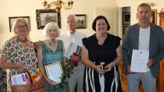 Fünf Menschen stehen lächelnd in einem Raum. Eine Frau hält ein gerahmtes Zertifikat und einen Korb. Eine andere Frau hält ein Buch. Hinter ihnen schmücken eine Kronleuchter und gerahmte Bilder die Wand.